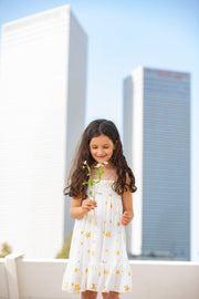 ילדה עם שיער שחור מתותל מחזיקה ענף של פרחי שדה בלבן וצהוב. לובשת שמלה לבנה מבד טריקו סימפוניה עם הדפס כוכבים בזהב. 