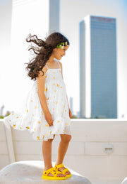 ילדה בשמלה לבנה מסתובבת מבד סימםוניה עם כוכבים בזהב. סנדלים בצבע צהוב ופרחי שדה בלבן צהוב בשיער. הילדה מצולמת מהצד והשמלה מתנפנפת עם הרוח אחורה